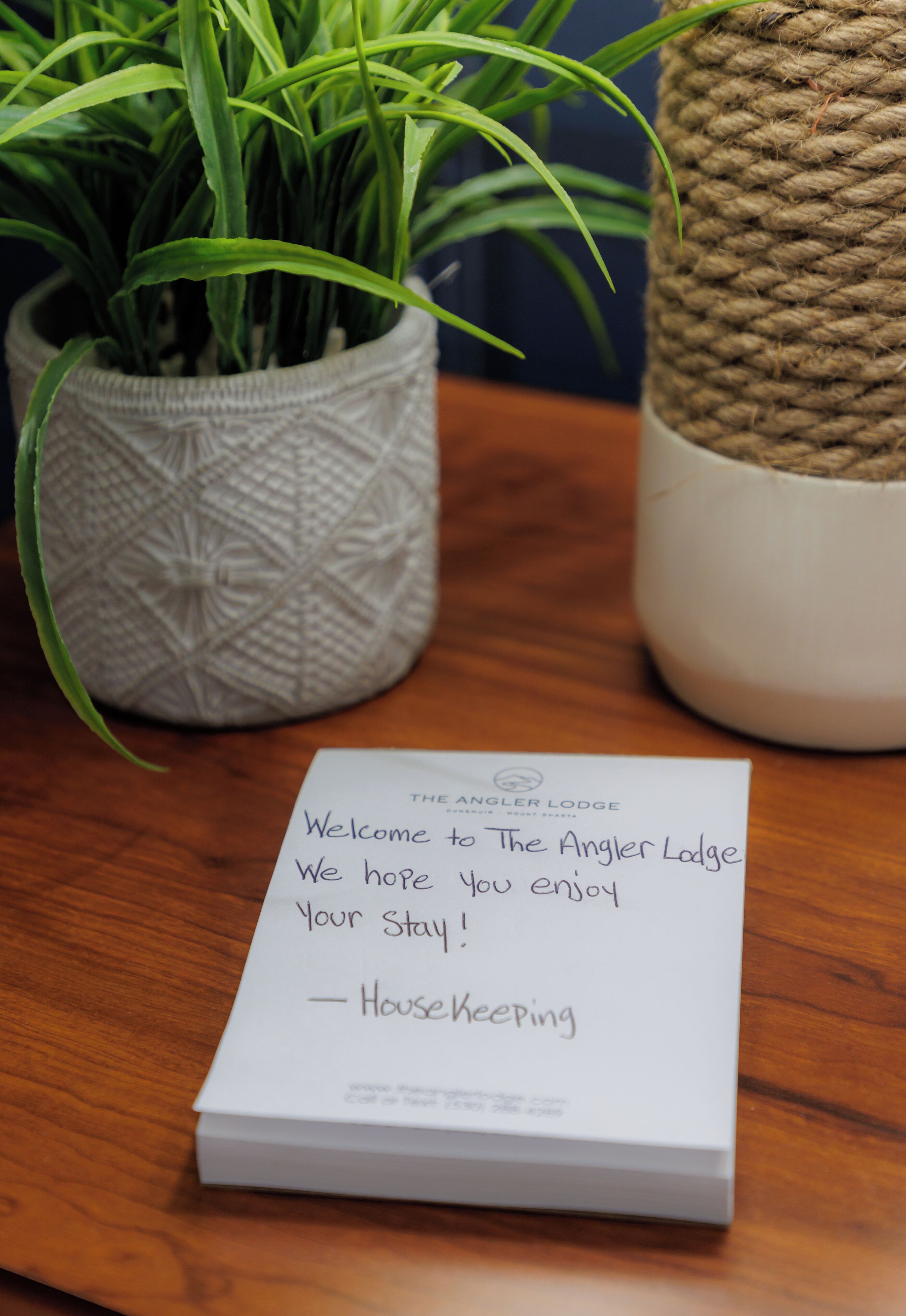 A welcome note from The Angler Lodge housekeeping, placed on a wooden table with a potted plant and a woven basket.