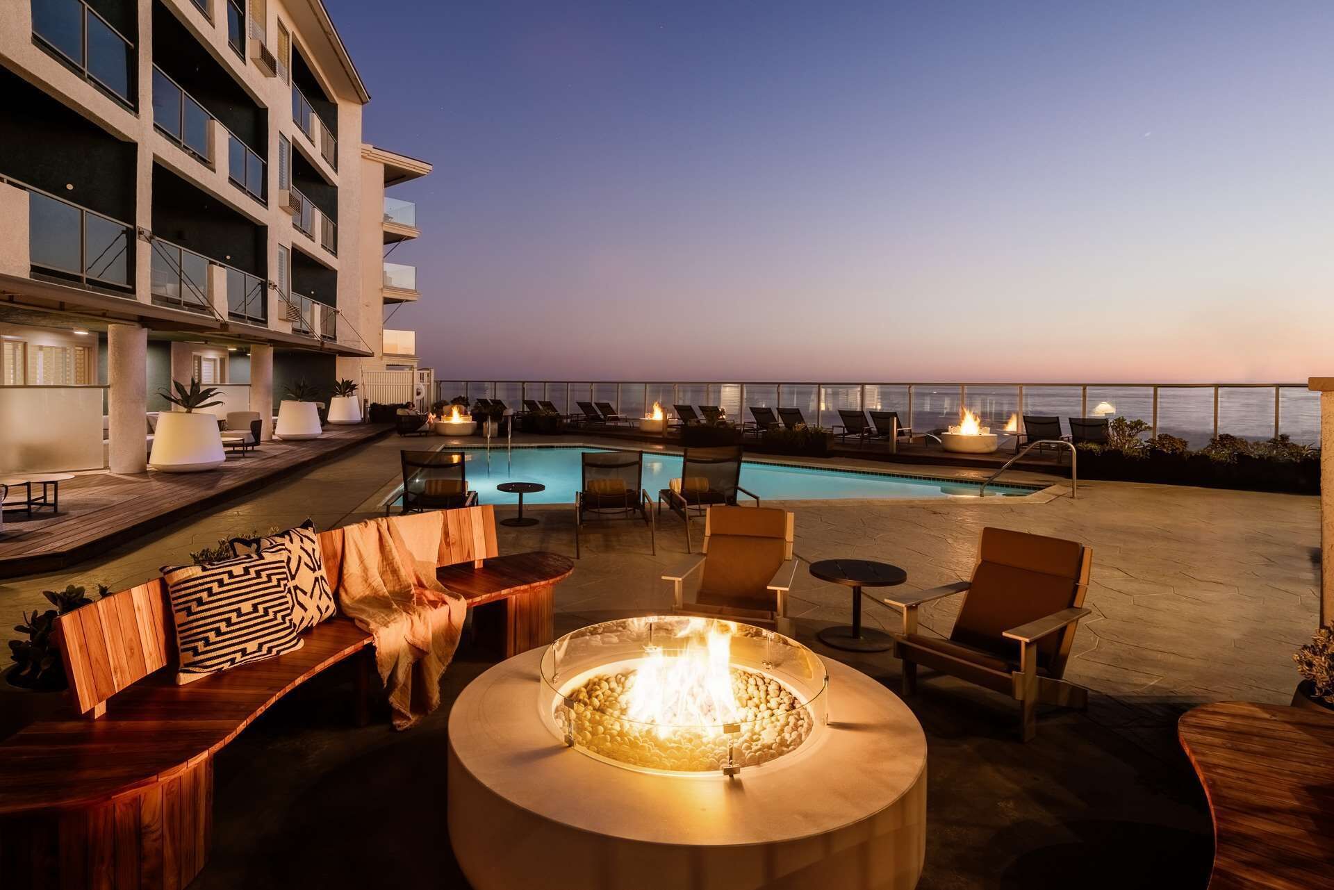 A rooftop pool area with lounge chairs, a fire pit, and a view of the ocean at dusk.