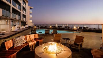 A rooftop pool area with lounge chairs, a fire pit, and a view of the ocean at dusk.