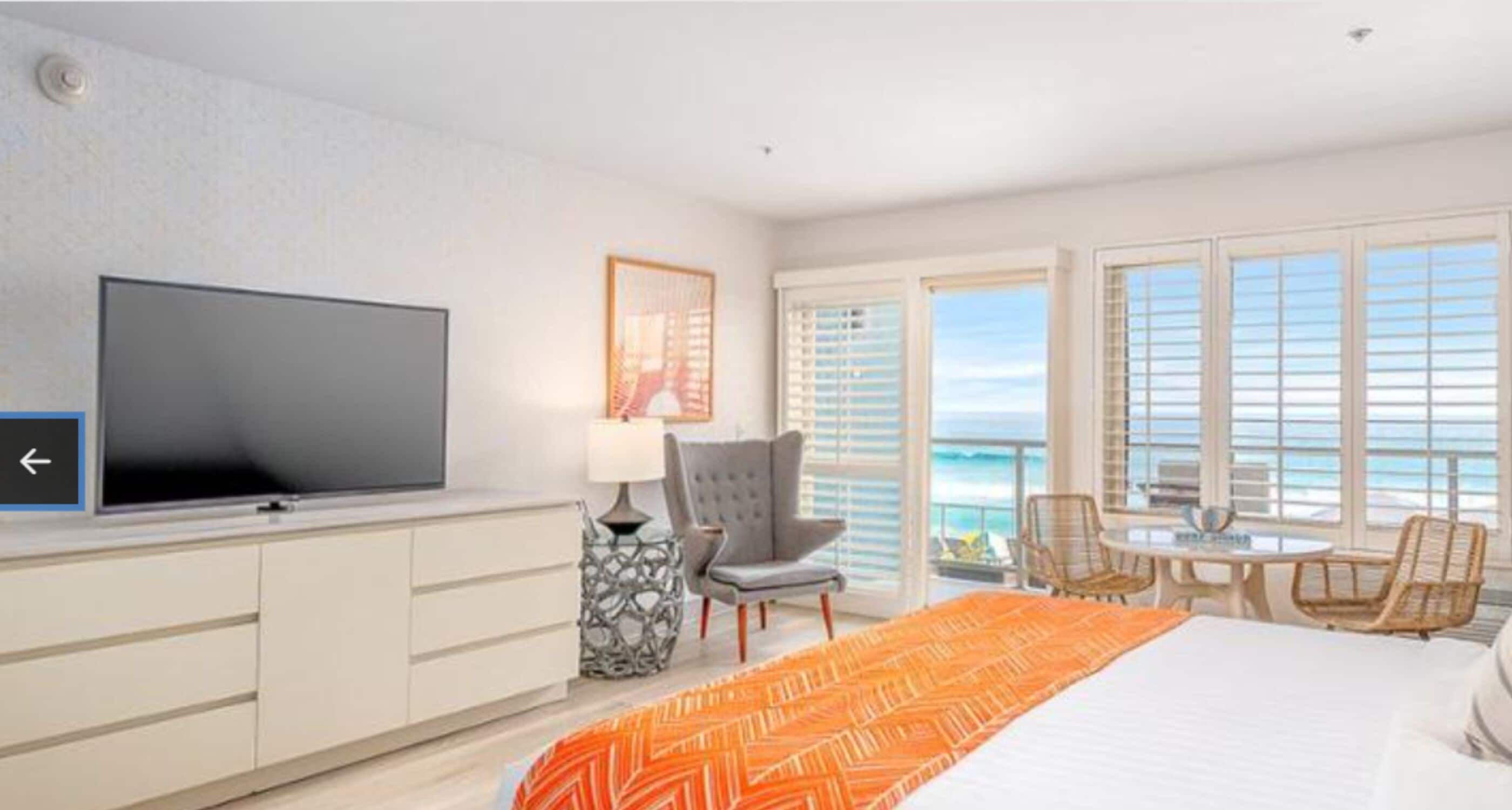 A hotel room with a bed, a TV, a desk with a chair, and a view of the ocean through large windows.