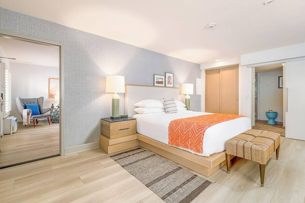 A modern hotel room with a large bed, bedside tables, a bench, and a view of the hallway.