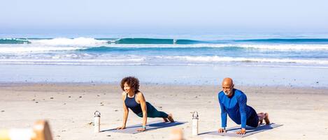 Duas pessoas praticando yoga em uma praia, com o oceano ao fundo.