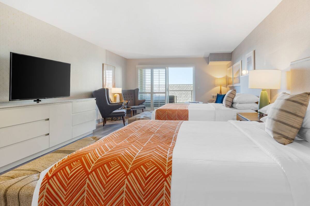 A hotel room with two beds, a TV, a desk, and a window with a view.
