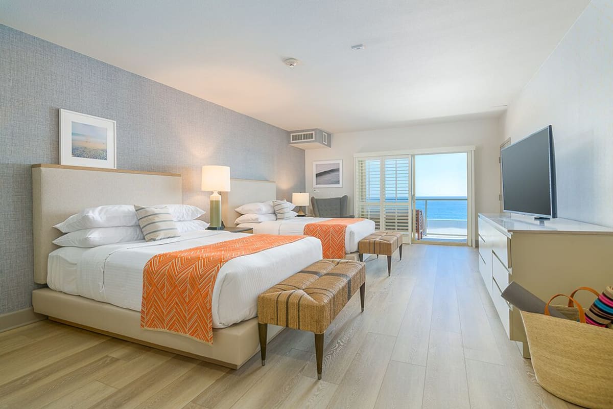 A hotel room with two beds, a television, a desk, and a view of the sea.