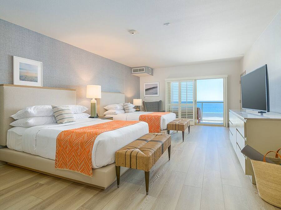 A hotel room with two beds, a television, a desk, and a view of the sea.