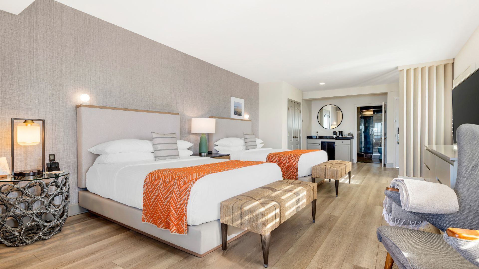 A hotel room with two beds, a sitting area, a small kitchenette, and a bathroom visible through a glass door.