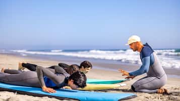 Escena de playa con tres personas acostadas sobre tablas de surf, una persona de pie gesticulando, y una vista despejada del océano al fondo.