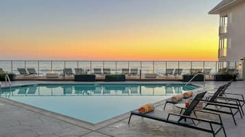 Zona de piscina con sillones reclinables y vista al océano al atardecer.
