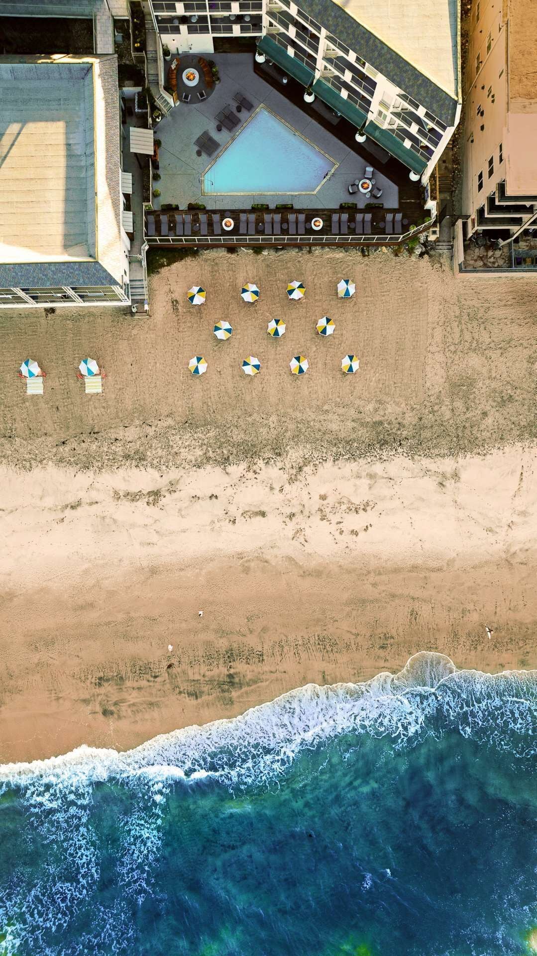Ein Luftbild zeigt einen Strand mit einem Poolbereich, einschließlich Liegestühlen und Sonnenschirmen.