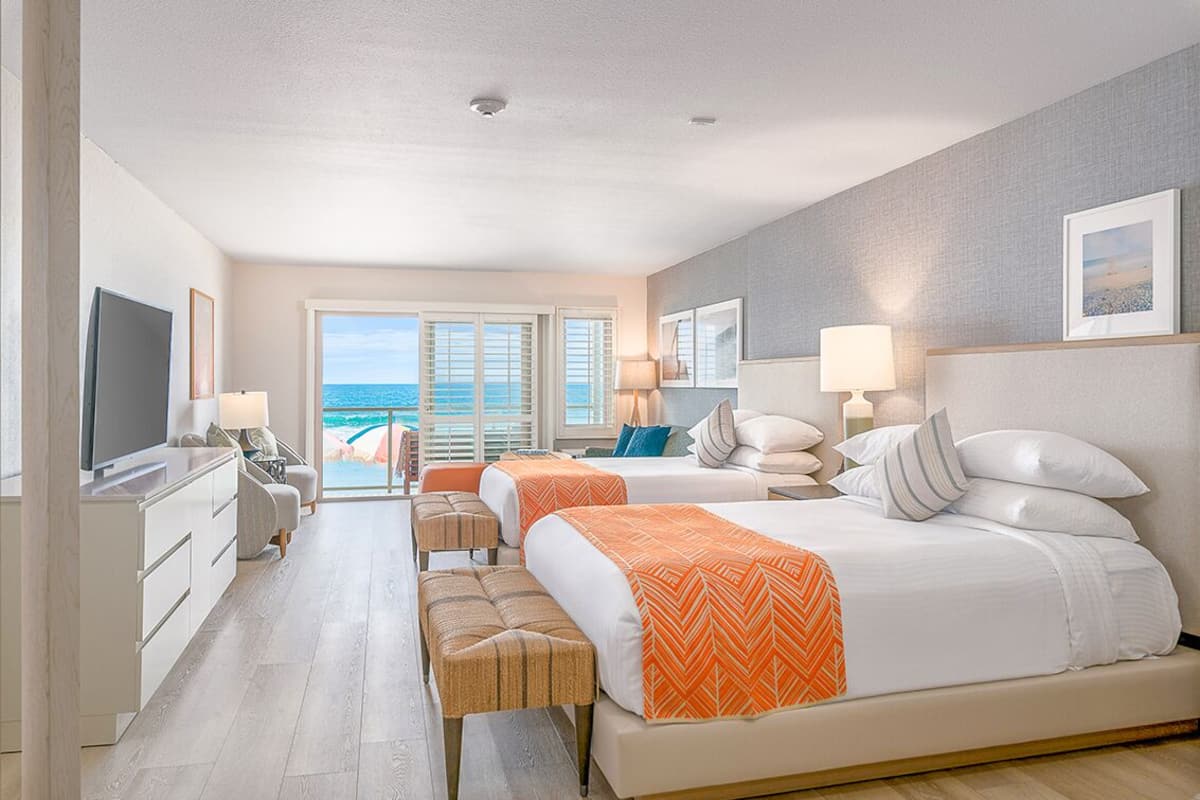 A hotel room with a large bed, two bedside tables, a TV, a desk, and a view of the ocean through large windows.