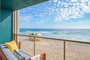 A balcony with a view of the beach and ocean, featuring a sofa with cushions and a table with an umbrella.