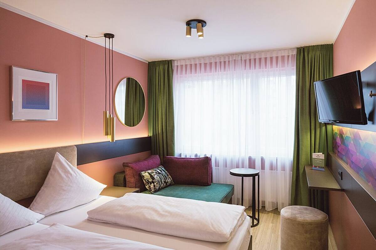 A hotel room with two beds, a desk, a television, a mirror, and a window with sheer curtains.