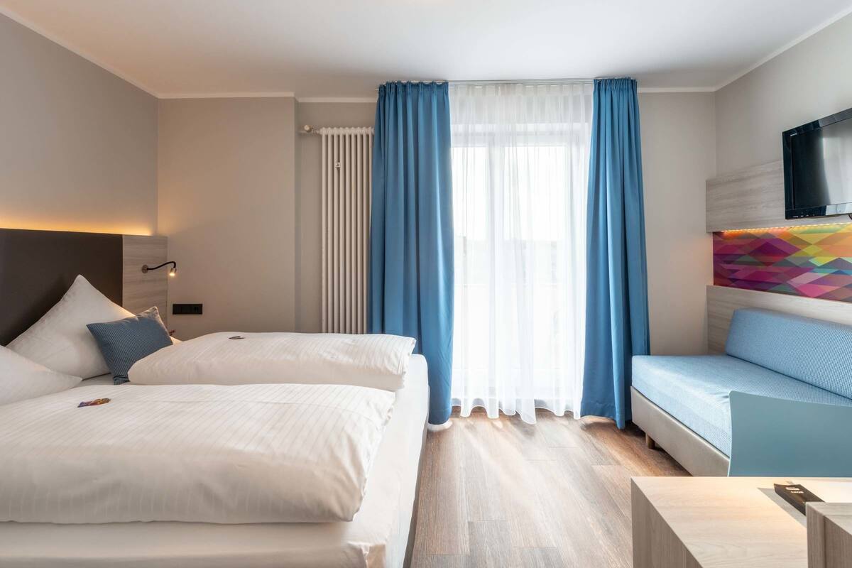 A hotel room with two beds, a sofa, a TV, and a colorful wall decoration.
