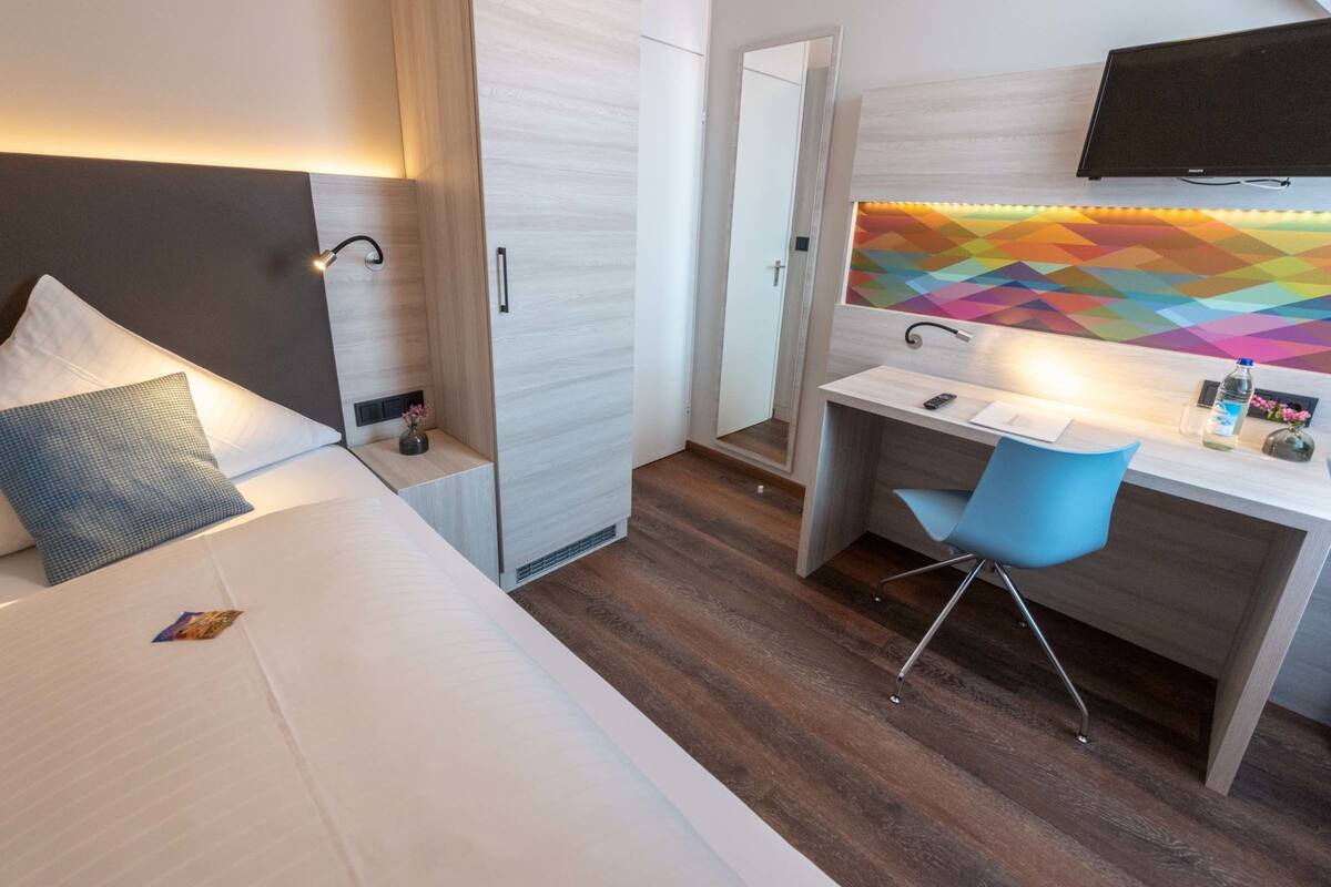 A hotel room with a bed, a desk with a chair, a mirror, and a colorful geometric wall decoration.