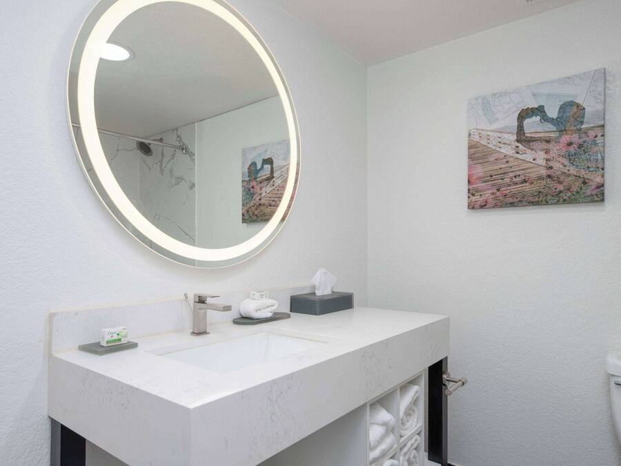 Ett badrum med ett vitt handfat, en rund spegel med en lampa, en tavla på väggen och en toalett.