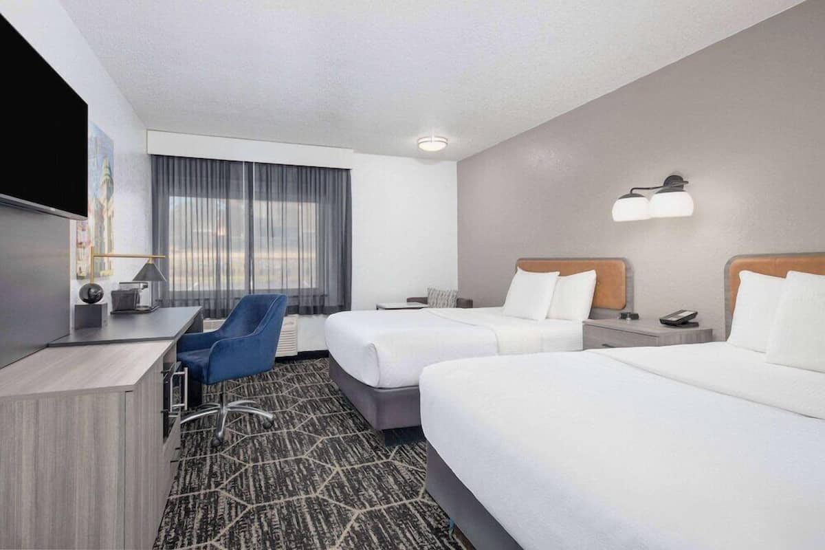 A hotel room with two beds, a desk, a chair, a TV, and a large window with curtains.