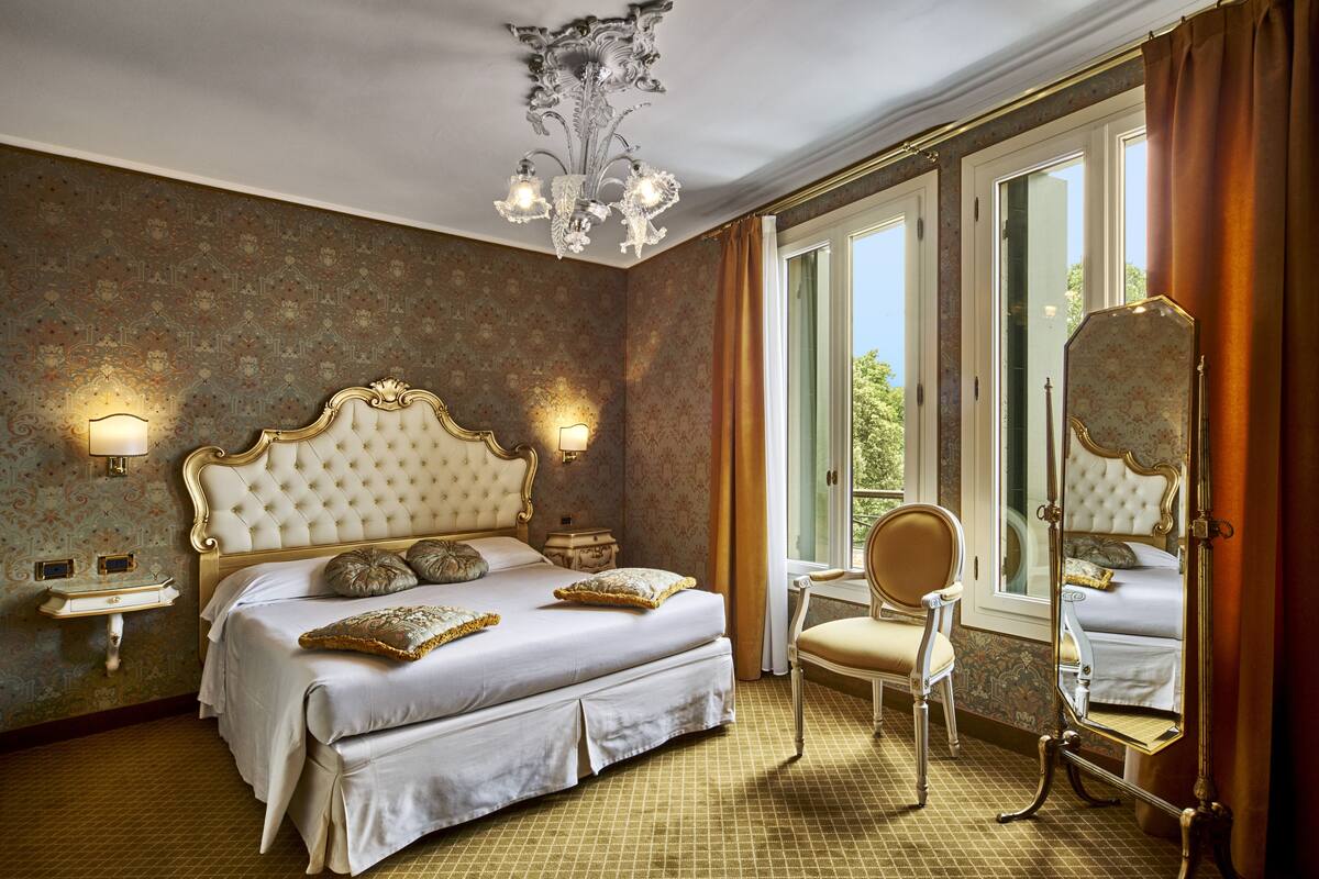 Un dormitorio lujoso con una cama grande, un cabecero decorado, dos mesitas de noche, un candelabro, un espejo y vistas al exterior.