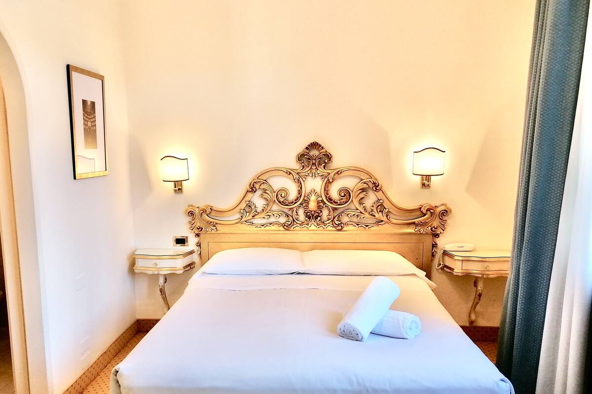 Una cama bien hecha con edredón blanco y almohadas, dos mesitas de noche con lámparas y un cabecero decorativo.