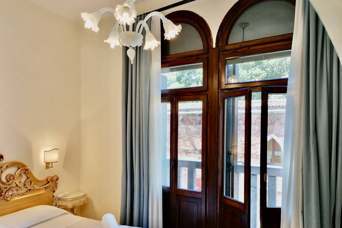 Habitación de hotel con cama, mesita de noche, teléfono, candelabro y vista al exterior.