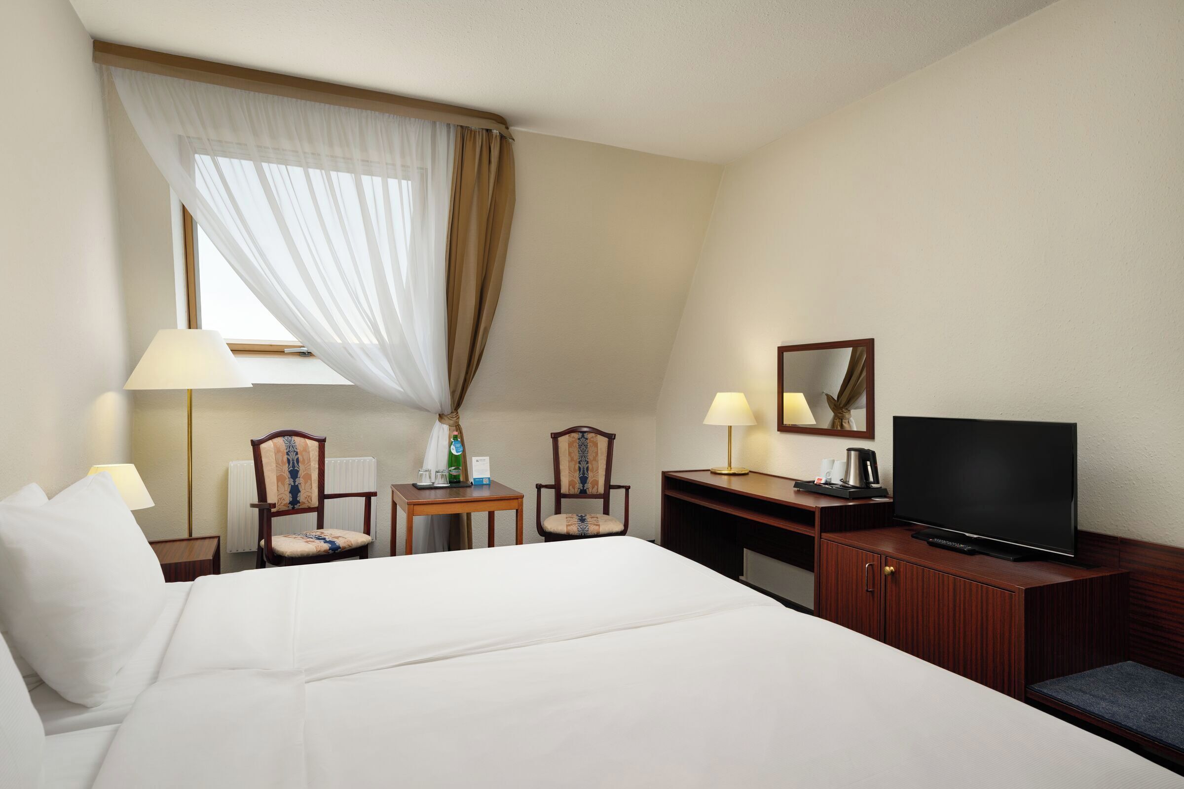 A hotel room with a large bed, two chairs, a desk with a television, and a window with sheer curtains.