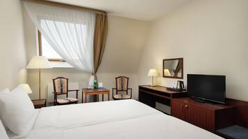 Habitación de hotel con una cama grande, dos sillas, un escritorio con televisor y una ventana con cortinas traslúcidas.