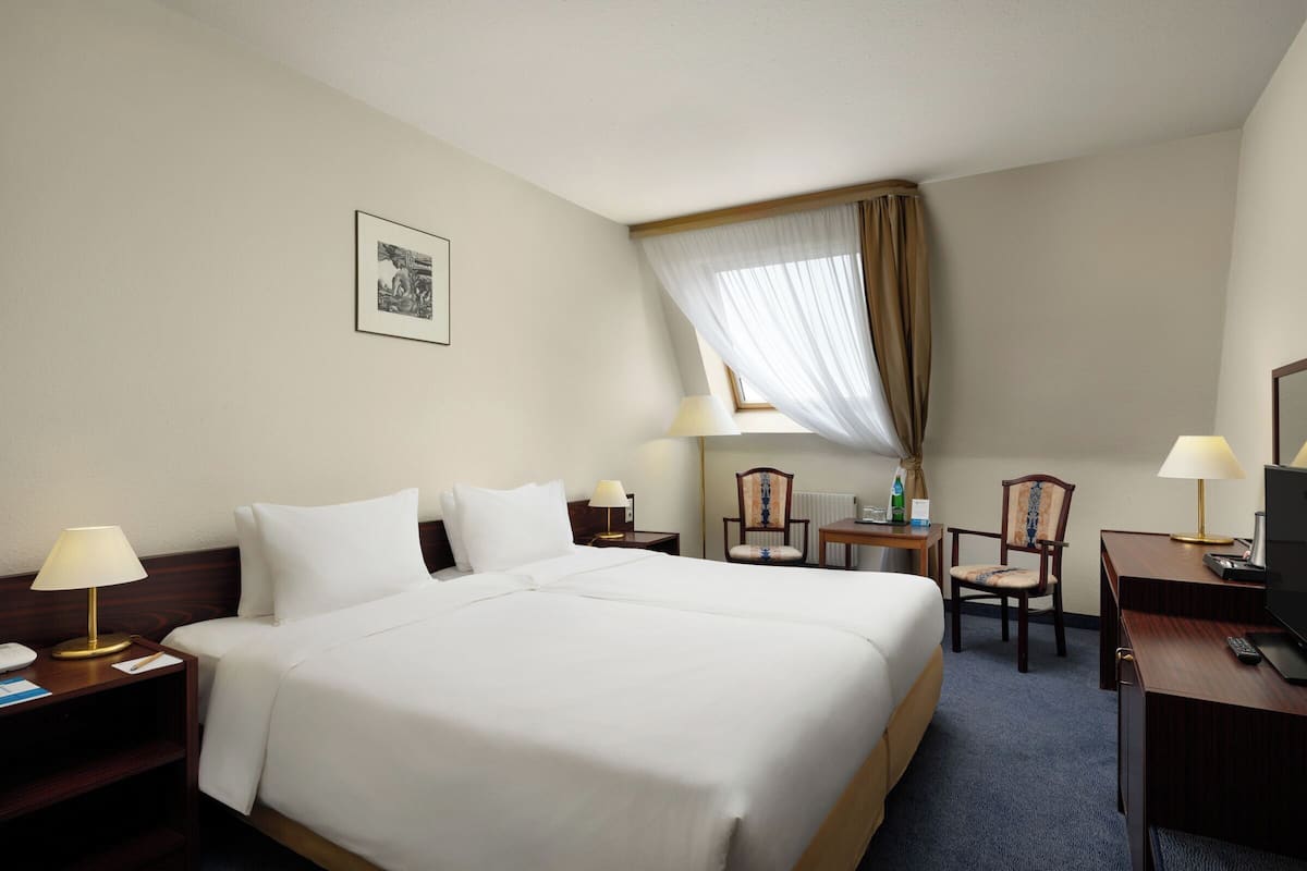 Habitación de hotel con una cama grande, dos mesitas de noche con lámparas, un escritorio con silla, un televisor y una ventana con cortinas.