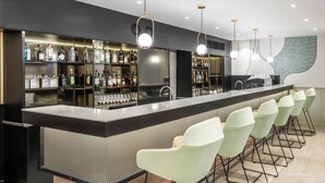 Un espace bar moderne, doté d’un long comptoir, de plusieurs suspensions lumineuses et d’un bar bien garni de diverses bouteilles. Plusieurs chaises vert clair sont disposées le long du comptoir.