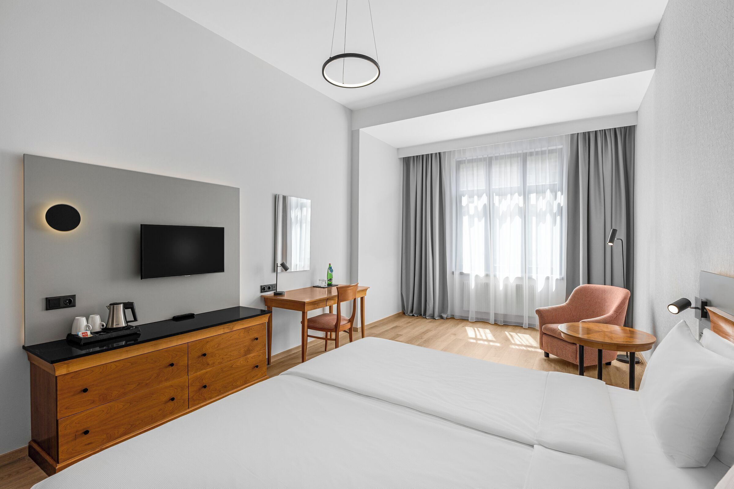 Una moderna camera d'albergo con un letto grande, una scrivania in legno, una televisione a schermo piatto, un tavolino, una sedia e un'ampia finestra con tende.