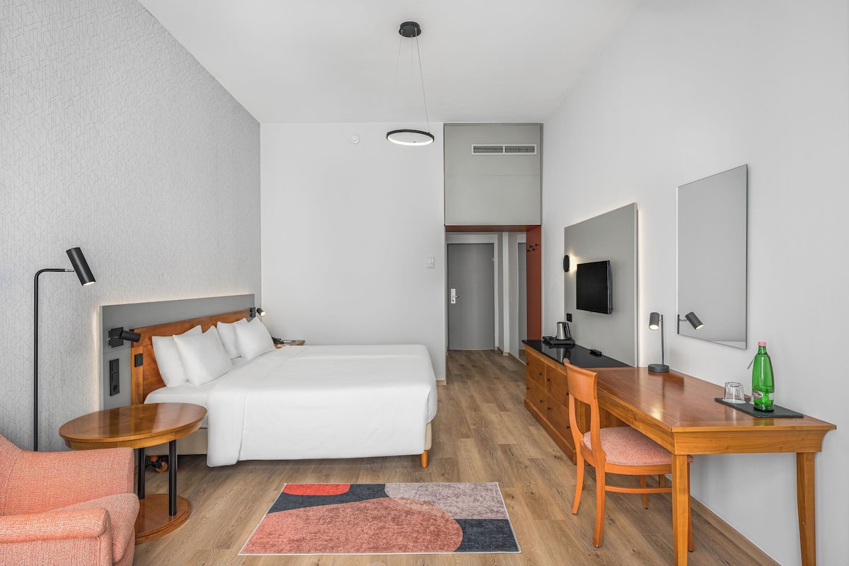 Habitación de hotel con cama, escritorio, silla, televisor y espejo.