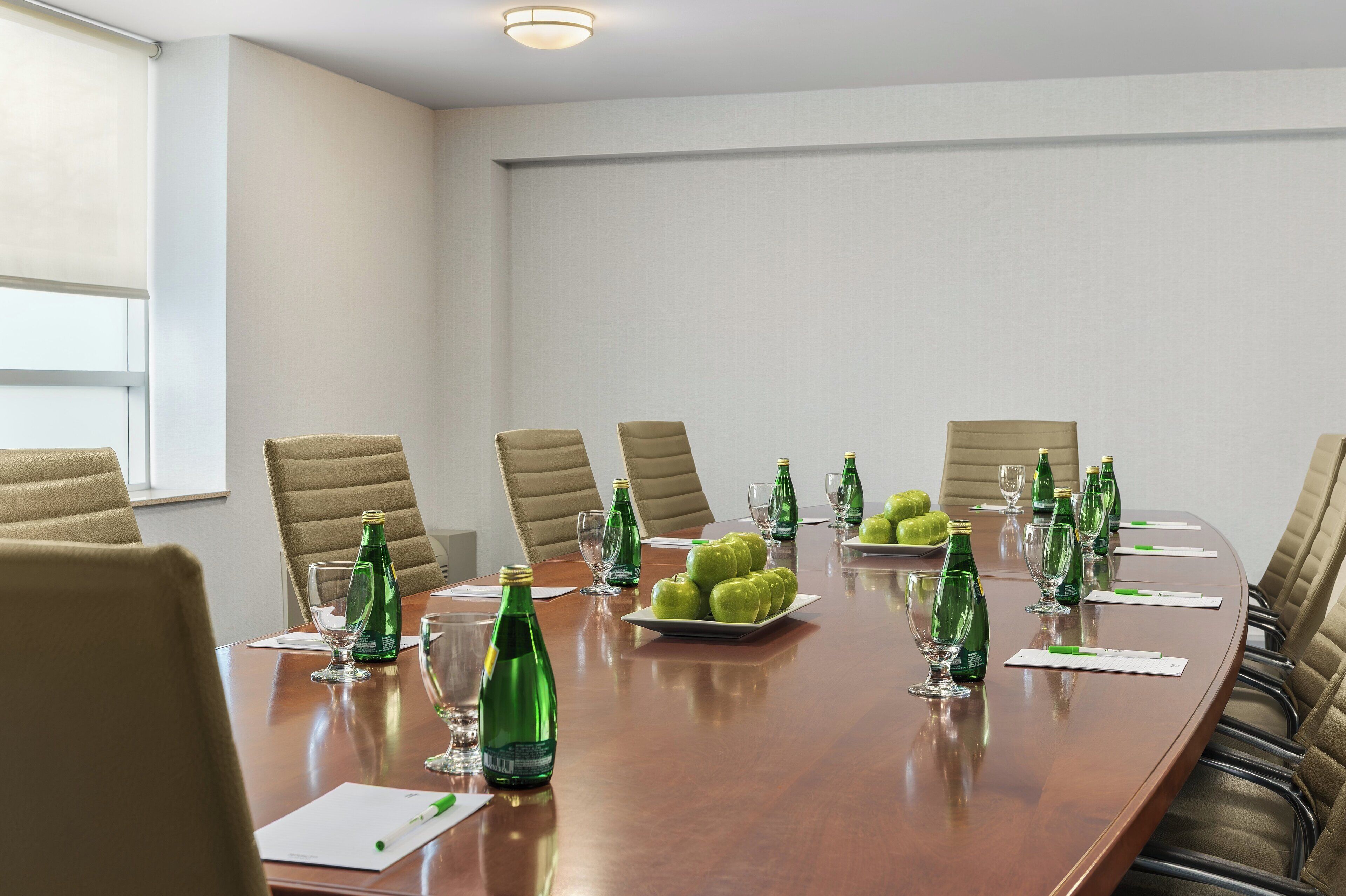A conference room with a long table, beige chairs, green bottles, glasses, and a plate of green apples.