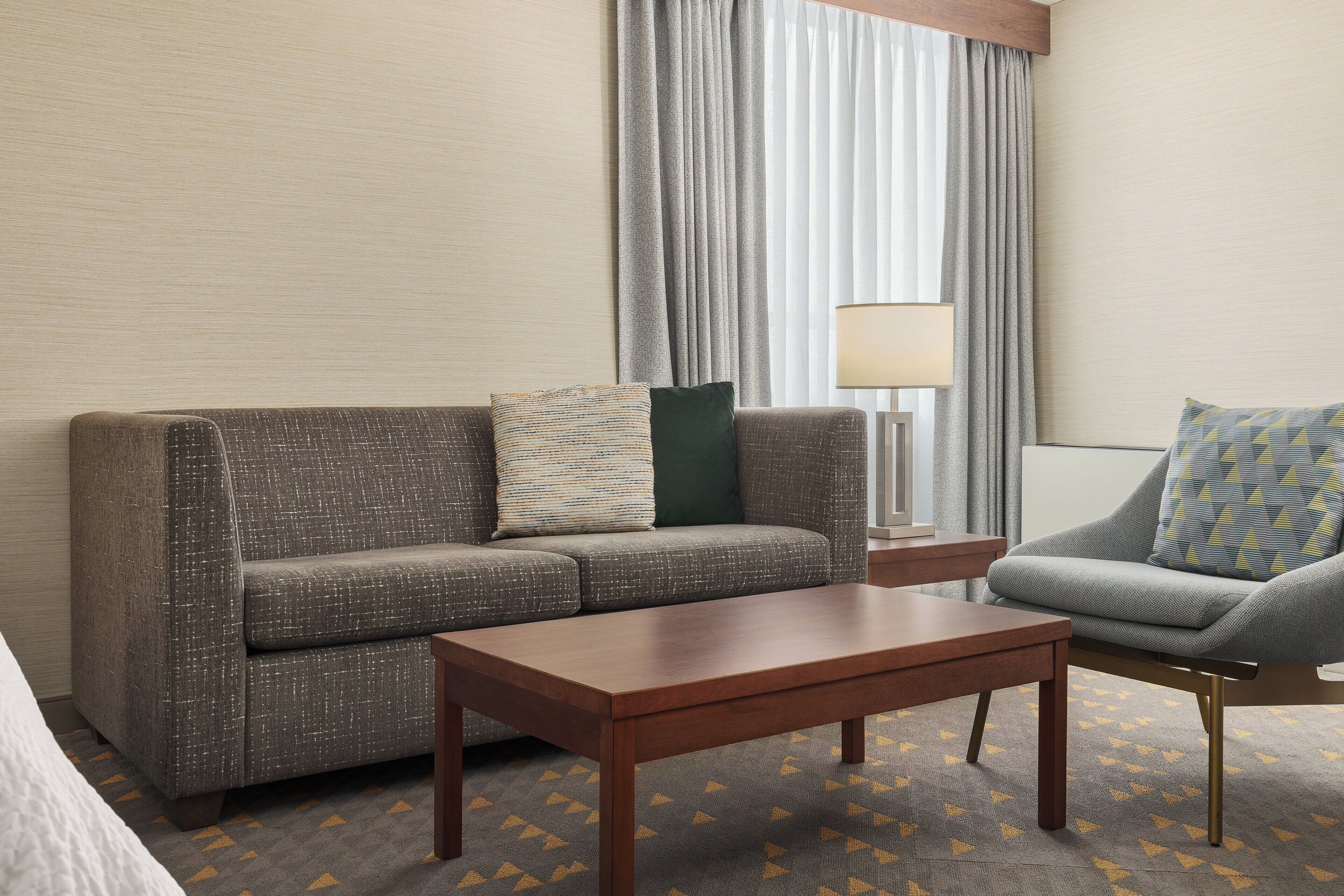 A hotel room with a grey sofa, a matching armchair, a wooden coffee table, and a lamp.