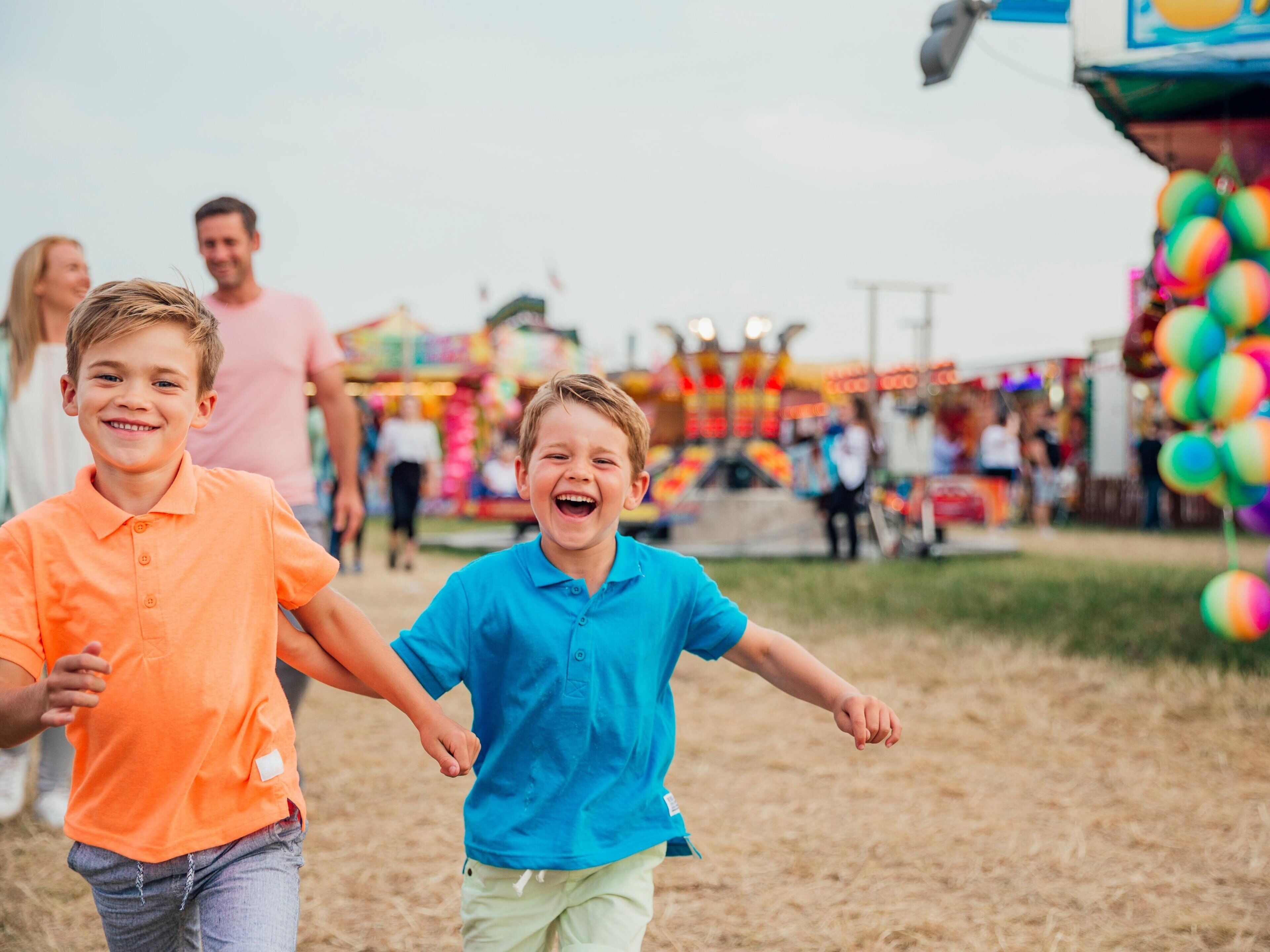 Two children holding hands in an outdoor setting with a fairground in the background.