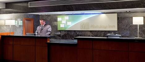 A reception desk at Holiday Inn with a marble wall backdrop, a man standing behind it, and a green sign with the hotel name.