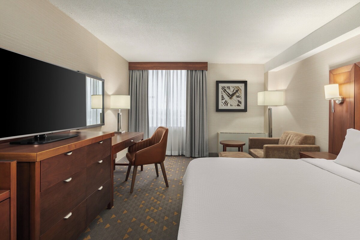 A hotel room with a bed, a desk with a TV, a chair, a lamp, and a framed picture on the wall.