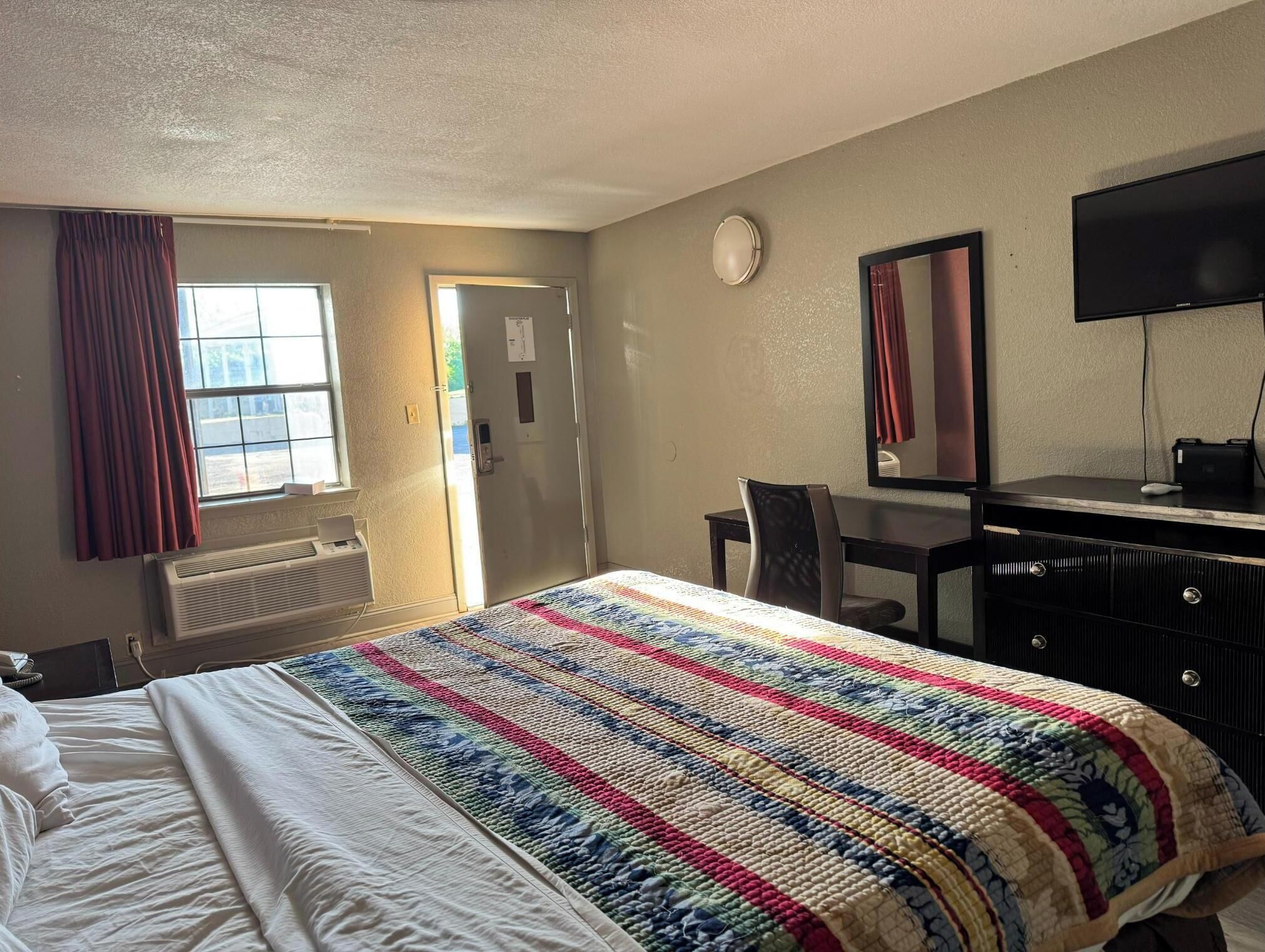 Una habitación de hotel con cama, escritorio, silla, televisor y ventana con cortinas.