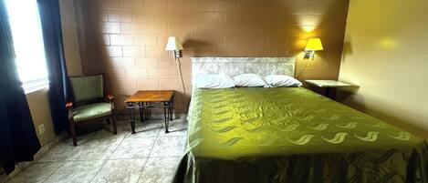 A hotel room with a bed, a chair, a small table, a lamp, and a window with curtains.