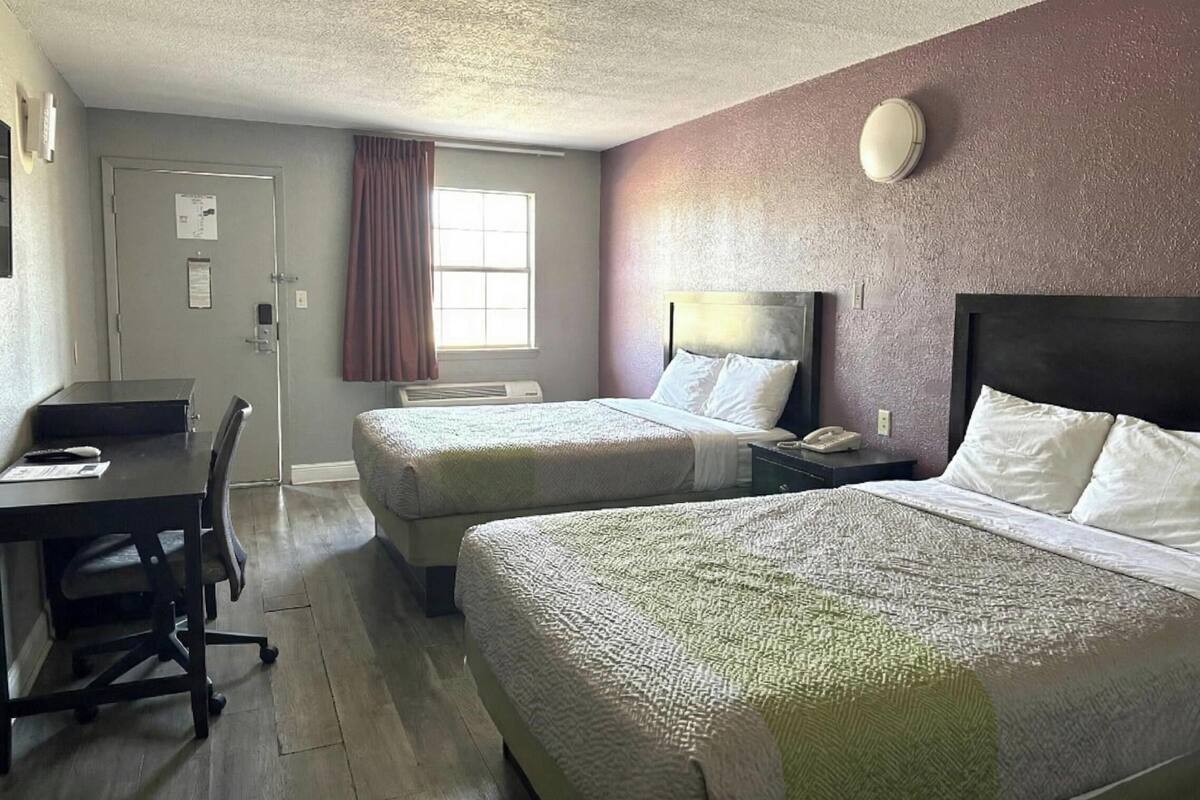 A hotel room with two beds, a desk, a chair, a window with curtains, and a door.