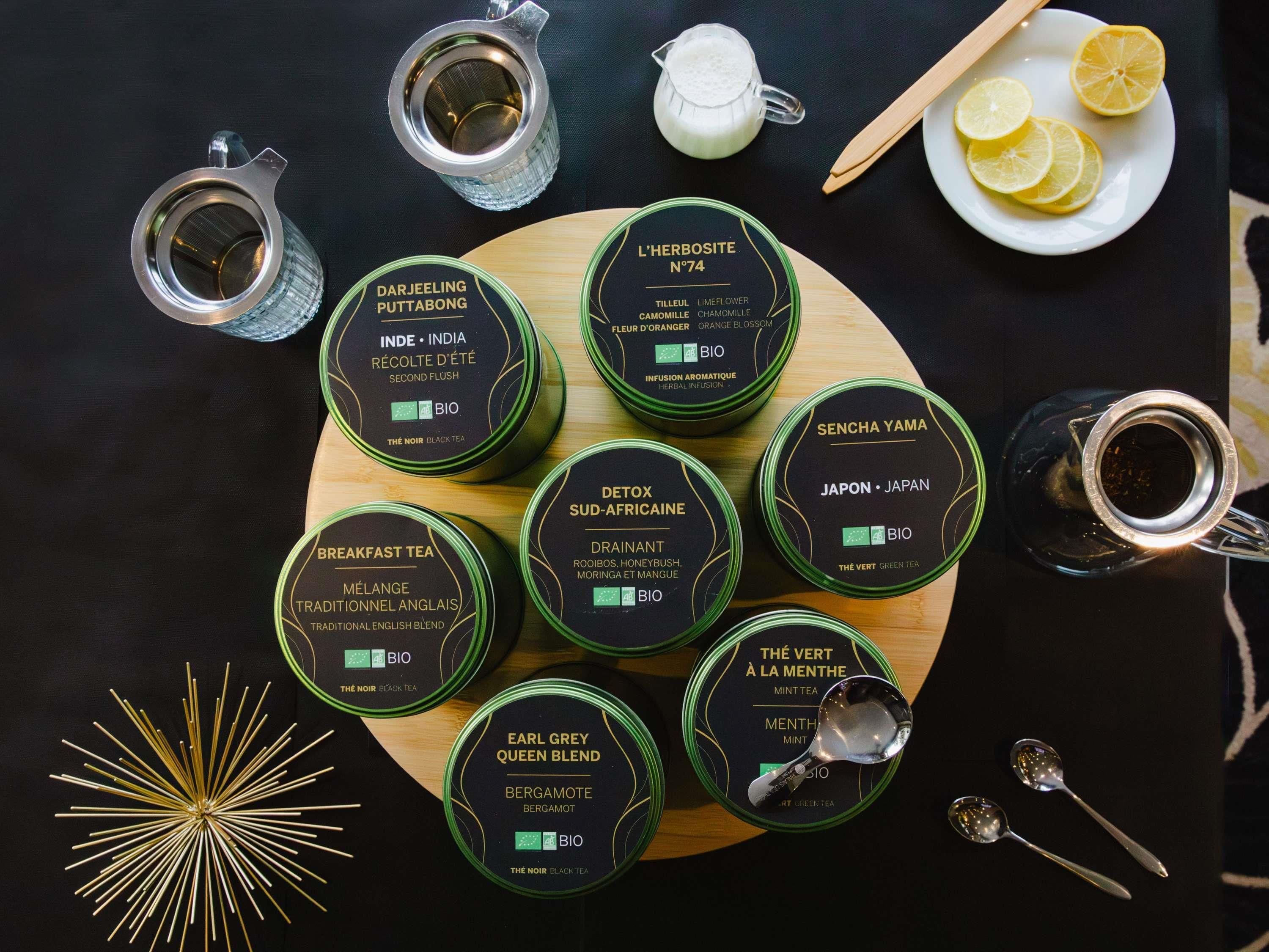 A variety of tea tins and a teapot on a wooden board, with a plate of lemon slices and a spoon set nearby.