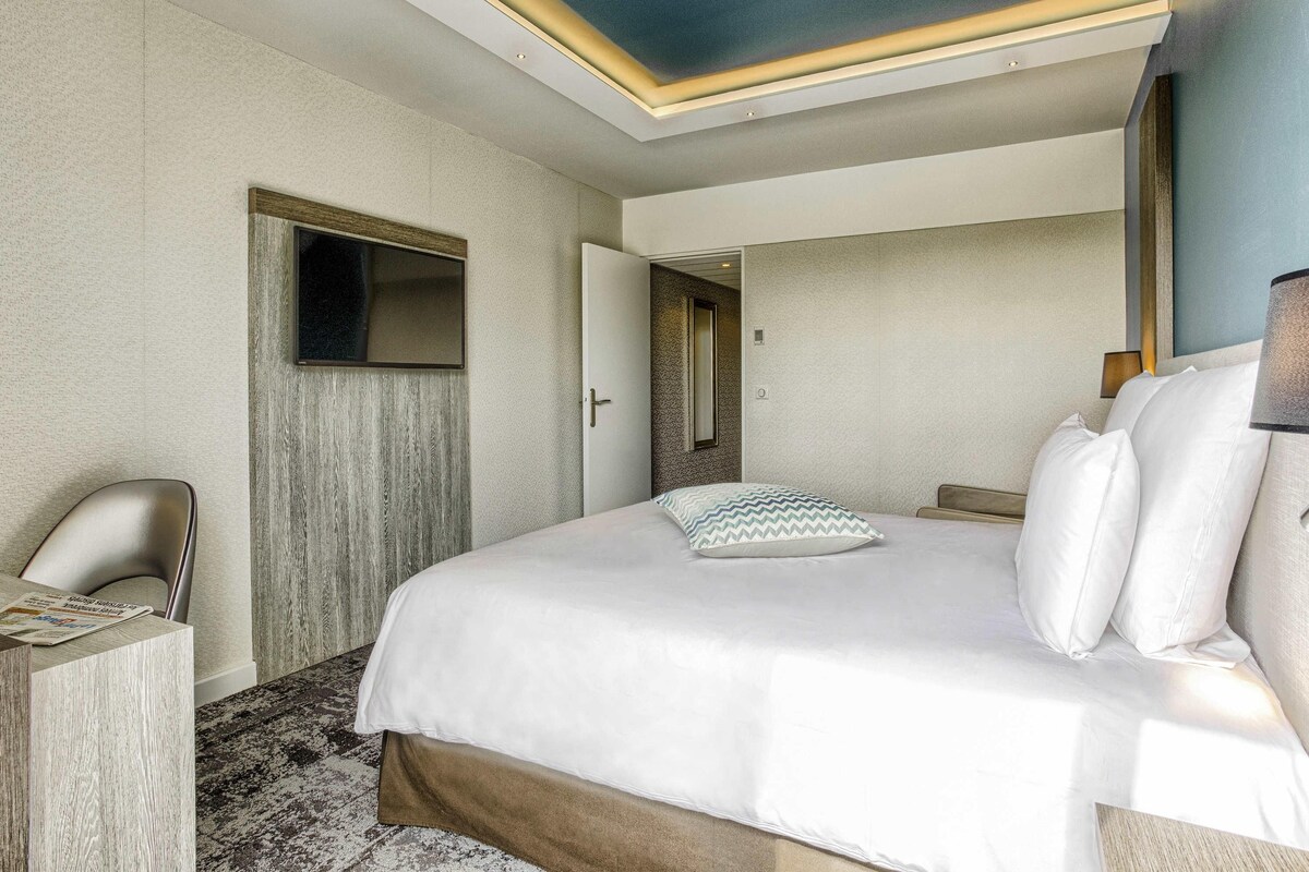 A hotel room with a bed, a desk, a chair, a mirror, and a door.