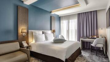 A hotel room with a bed, a desk, a chair, a sofa, and a large window with curtains.