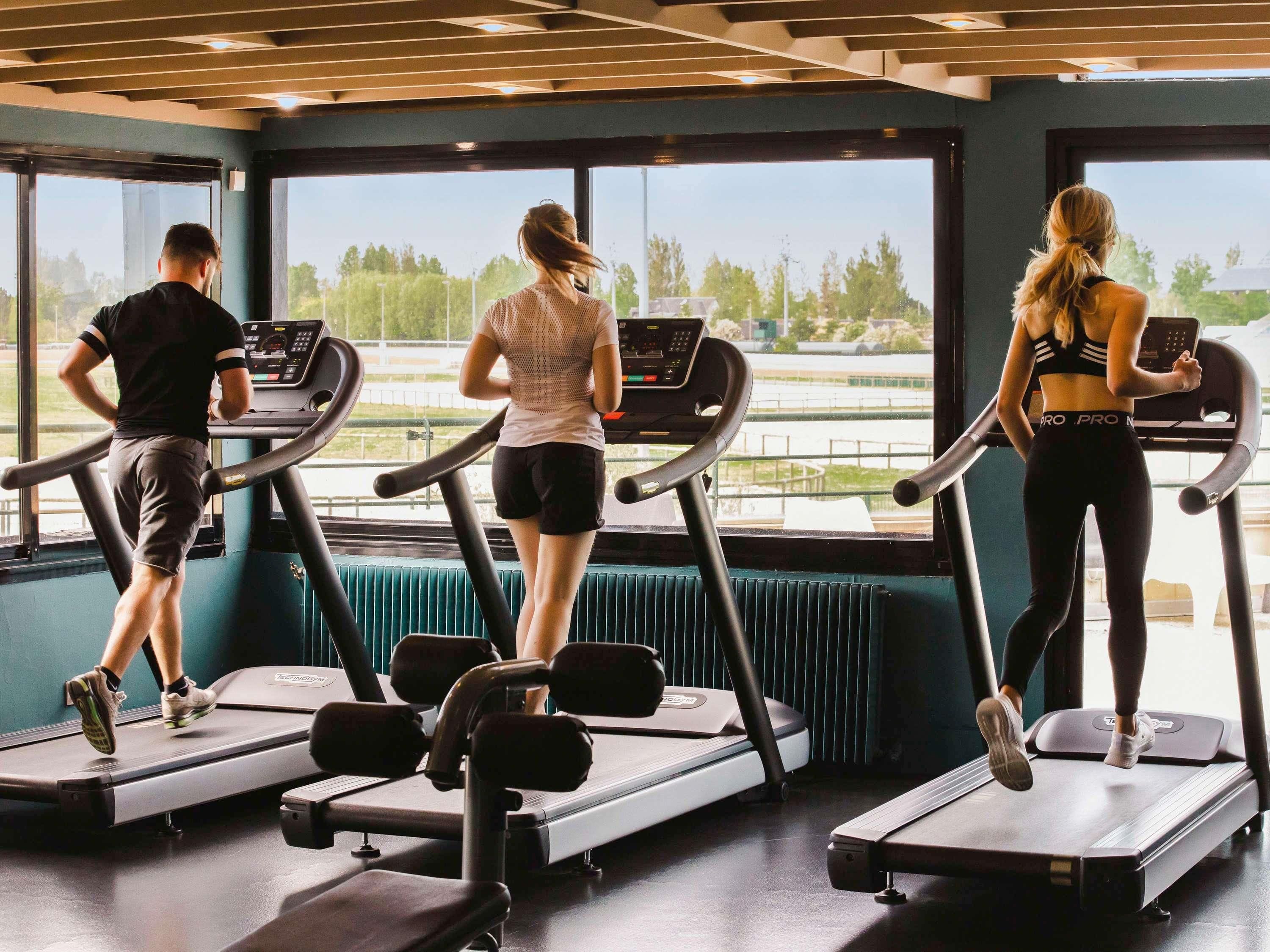 Trois personnes courent sur des tapis de course dans une salle de sport dotée de grandes fenêtres, offrant une vue sur une route et des arbres.