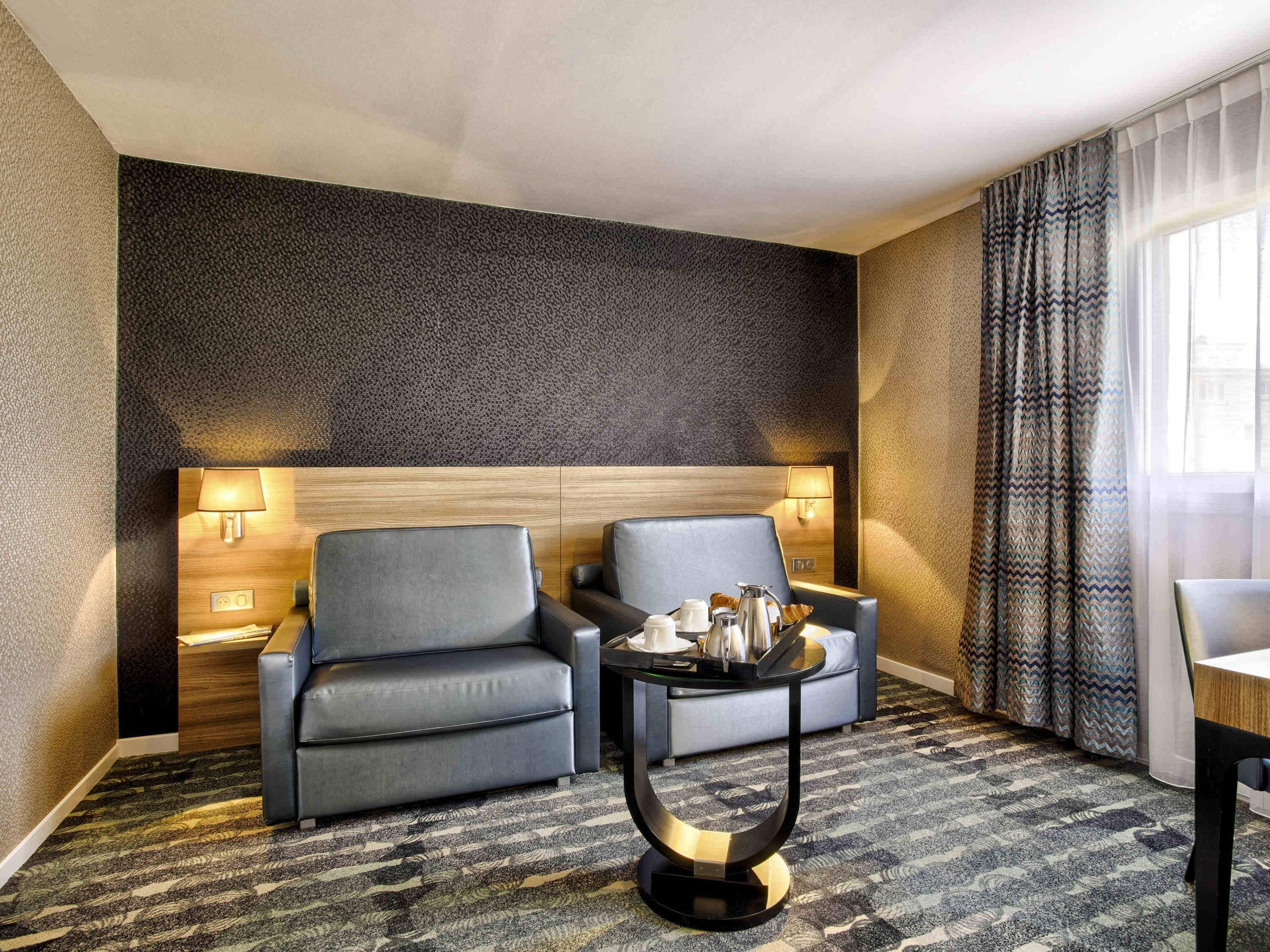 Une chambre d’hôtel avec un mur texturé de couleur sombre, deux fauteuils gris, une petite table ronde avec une lampe et un tapis à motifs.