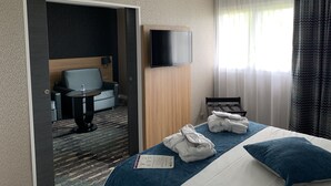 Habitación de hotel con cama, televisor montado en la pared, zona de estar con una silla y una mesita, y ventana con cortinas.