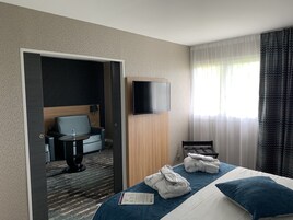 Habitación de hotel con cama, televisor montado en la pared, zona de estar con una silla y una mesita, y ventana con cortinas.