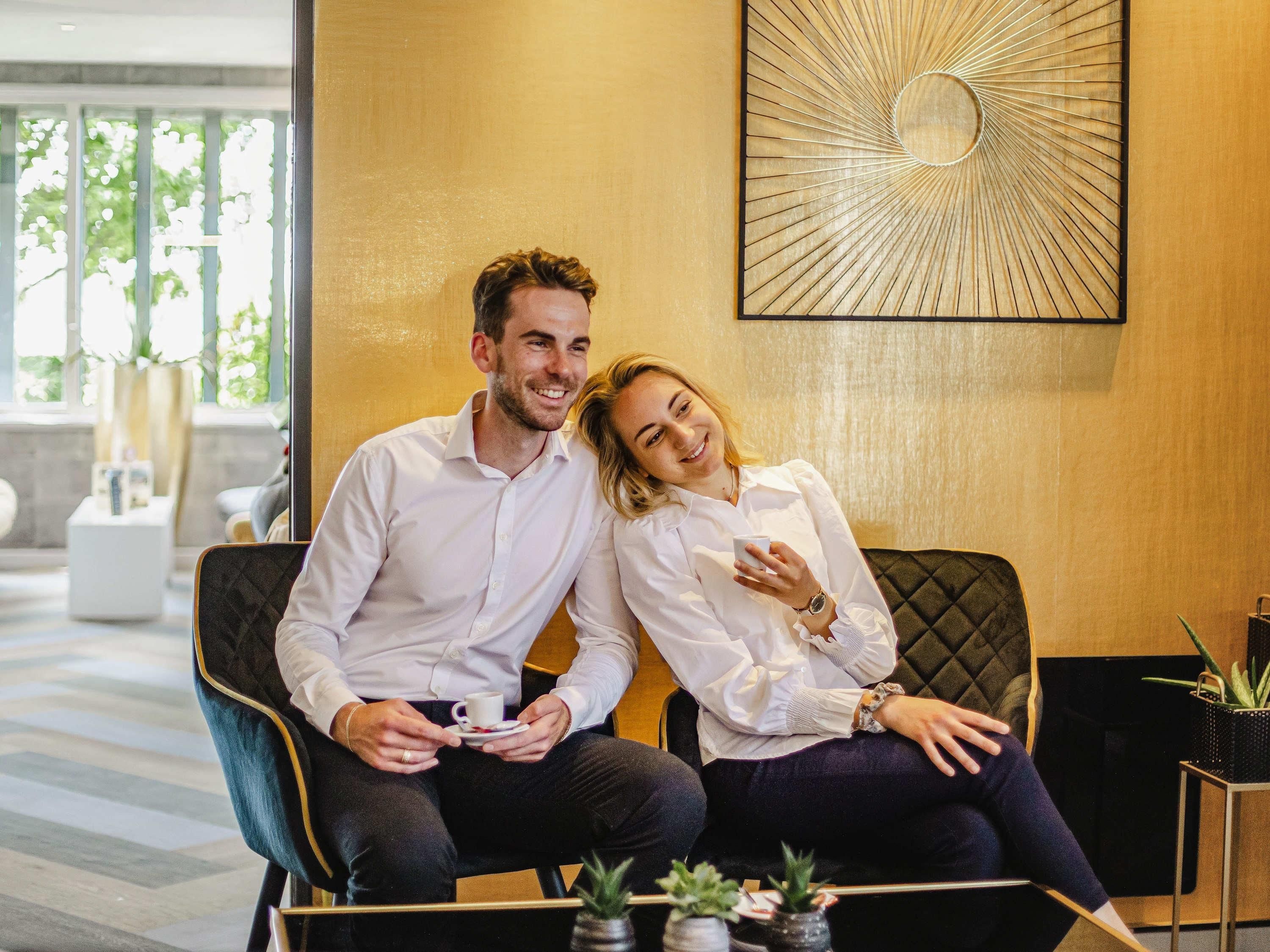 A couple sitting on a black leather couch in a modern lobby with a large abstract wall art piece above them.