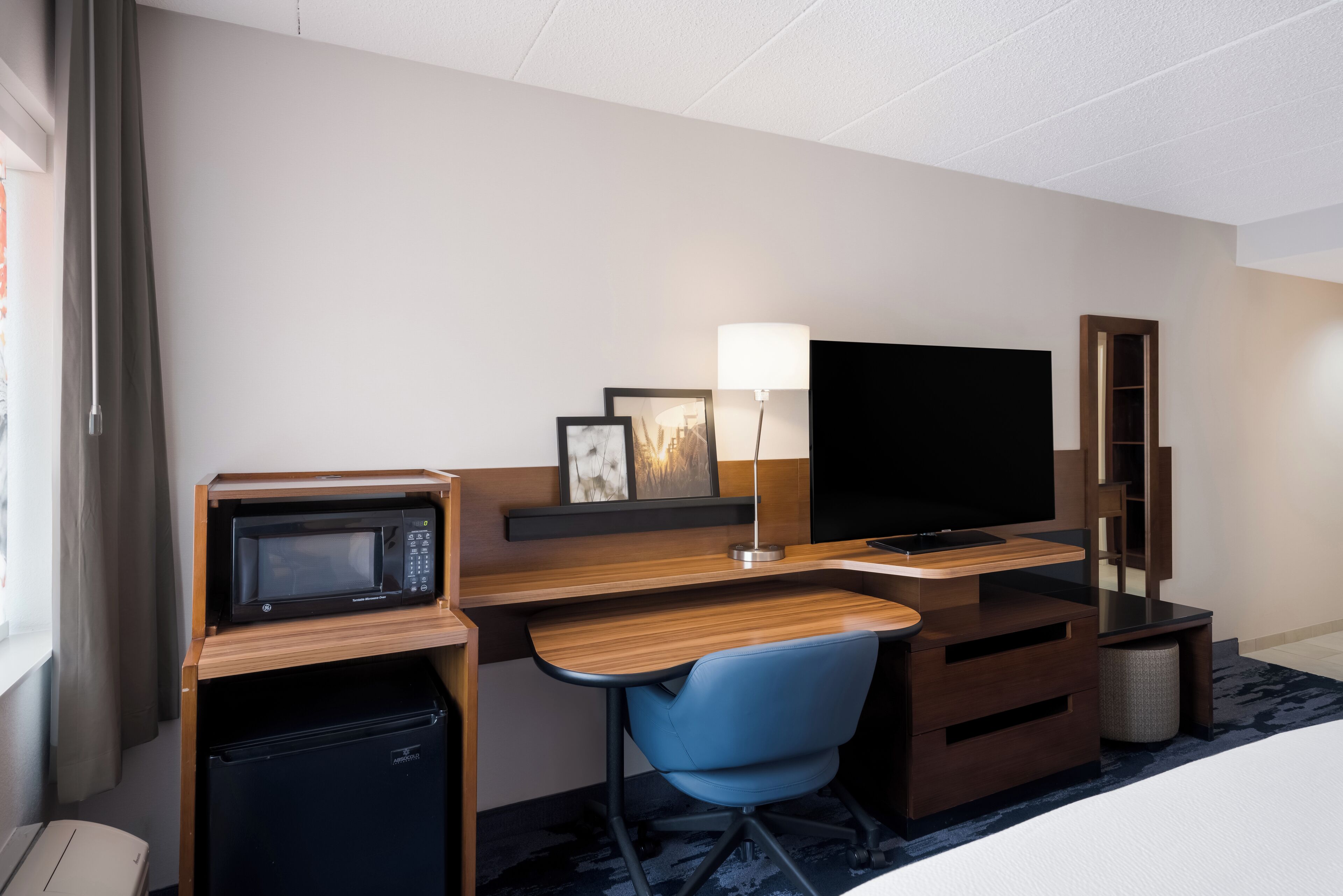 A hotel room with a wooden desk, a blue chair, a television, a microwave, and a small refrigerator.
