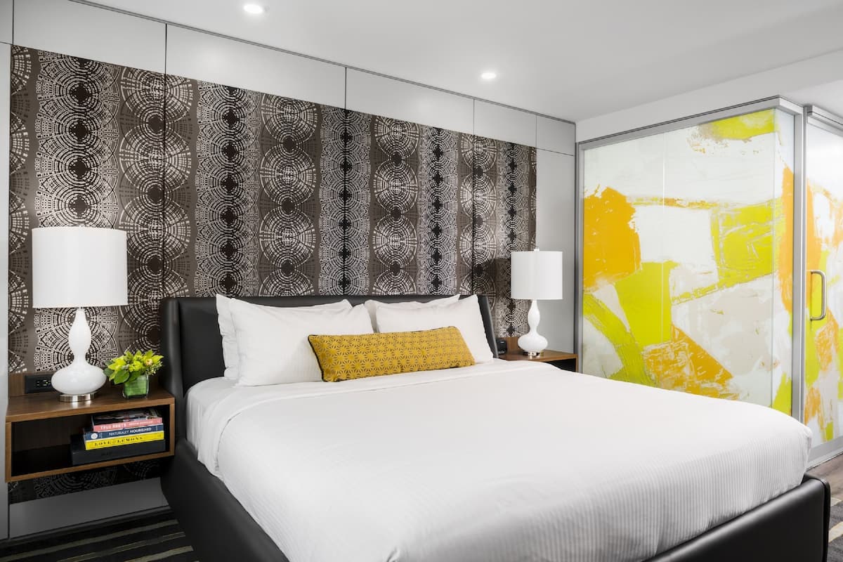 A modern hotel room with a large bed, two bedside tables with lamps, a patterned headboard, and a large abstract painting on the wall.