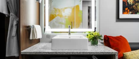 A modern bathroom with a granite countertop, a white sink, and a mirror. There is a towel rack with towels, a potted plant, and a cushion on the countertop.