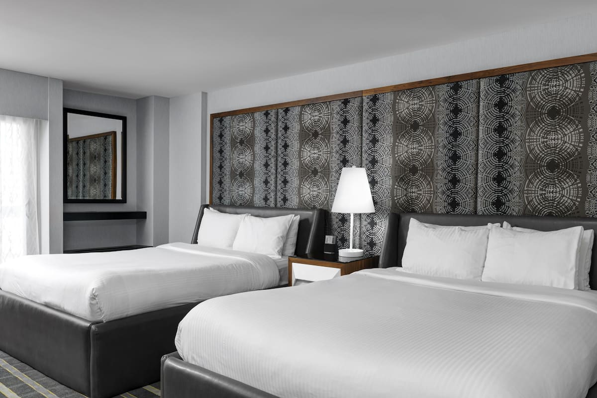 A hotel room with two beds, a large patterned headboard, a bedside table with a lamp, and a mirror on the wall.