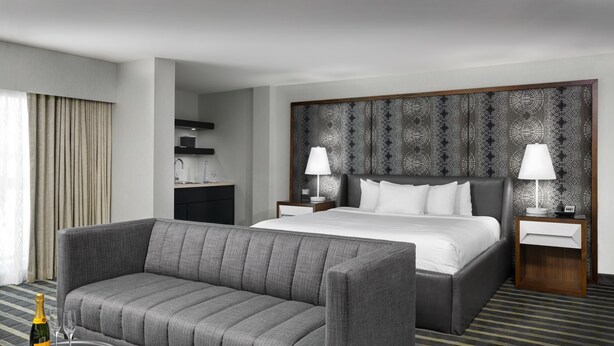 Une chambre d’hôtel moderne avec un canapé gris, un grand lit, une table de chevet avec une lampe et une petite table sur laquelle se trouvent une bouteille et des verres.